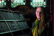 Photo of Catherine in front of stained-glass windows, next to a bookcase with leather-bound books. The 'Institute of Mining and Mechanical Engineers' logo is faintly visible below the window. She is wearing smart-casual clothes.