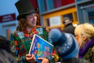 A photo of the Rock Showman, from the waist up holding a blue children's book titled 'Midnight Oil'. He wears a black top hat and multi colour tartan tails. He is stood talking with members of the public - adults and children, with their heads facing away from the camera in the foreground.