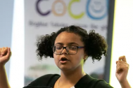 A photograph of a woman talking, with her hands raised. She stands in front of a banner with COCO's logo on.