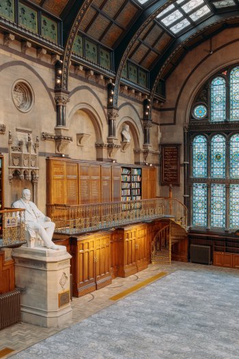 A photograph of The Common Room's Wood Hall featuring colourful stained glass windows, wooden bookcases along the walls and balconies (some are open, so you can see some of the books), a large portrait hanging from the wall, and a large white statue of a man in the centre of the room.